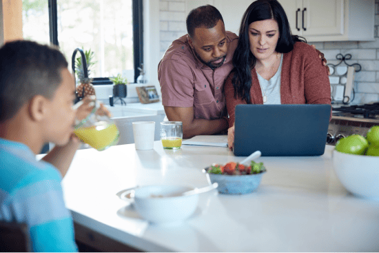 Couple looking at a laptop in a kitchen while a child drinks juice at the counter.